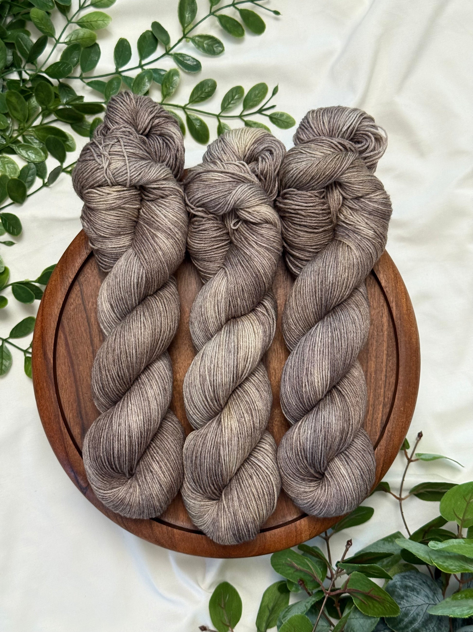 Three twisted hanks of heathered taupe-gray yarn arranged on a round wooden board, surrounded by green leaves on a white fabric background.