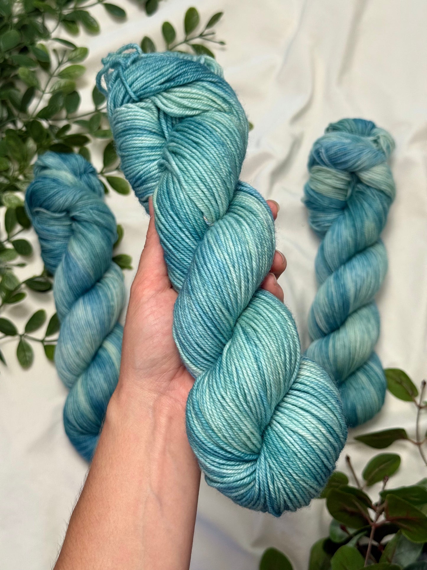 A hand holds a skein of variegated yarn in shades of teal and aqua, with two more skeins placed behind it on a white fabric background with green leaves around the edges.