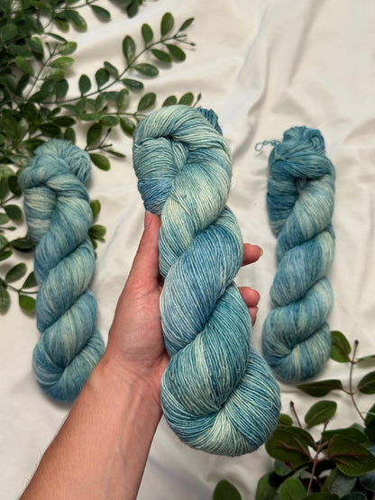 A hand holds a twisted hank of variegated blue and mint green yarn, with two more matching skeins placed behind it on a white fabric background with green leaves around the edges.