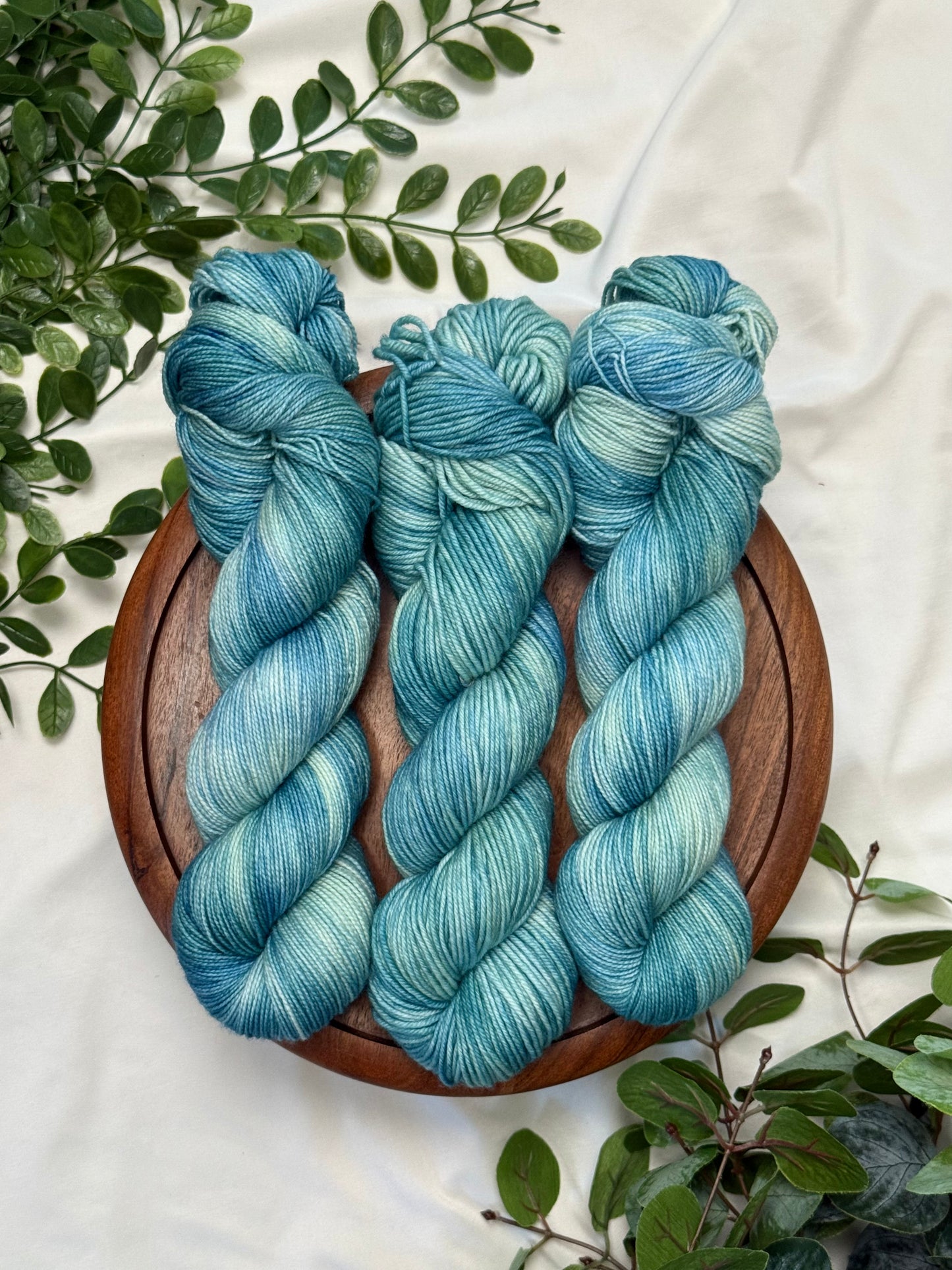 Three twisted hanks of variegated blue and teal yarn arranged on a round wooden tray, surrounded by green foliage on a white fabric background.