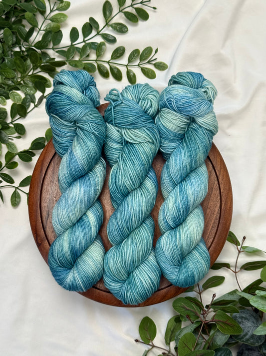Three twisted hanks of variegated blue and teal yarn arranged on a round wooden tray, surrounded by green foliage on a white fabric background.