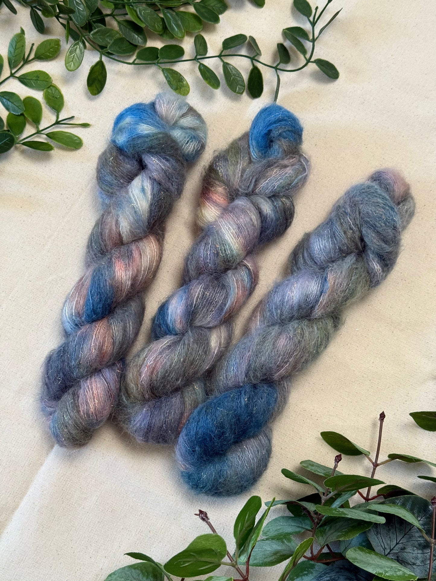 Three hanks of variegated alpaca yarn in shades of blue, gray, and soft pink, arranged on a beige fabric background with green foliage accents.
