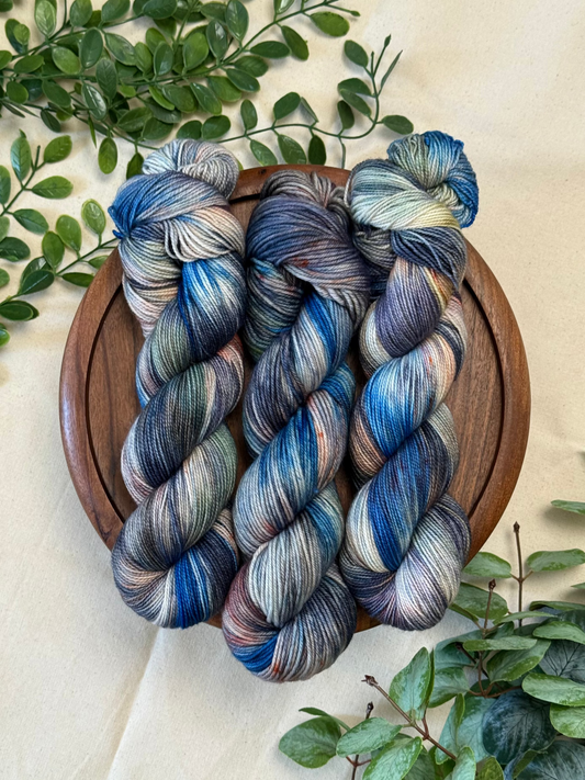 Three hanks of variegated yarn in shades of blue, gray, purple, and cream, arranged on a round wooden tray with green foliage accents on a beige fabric background.