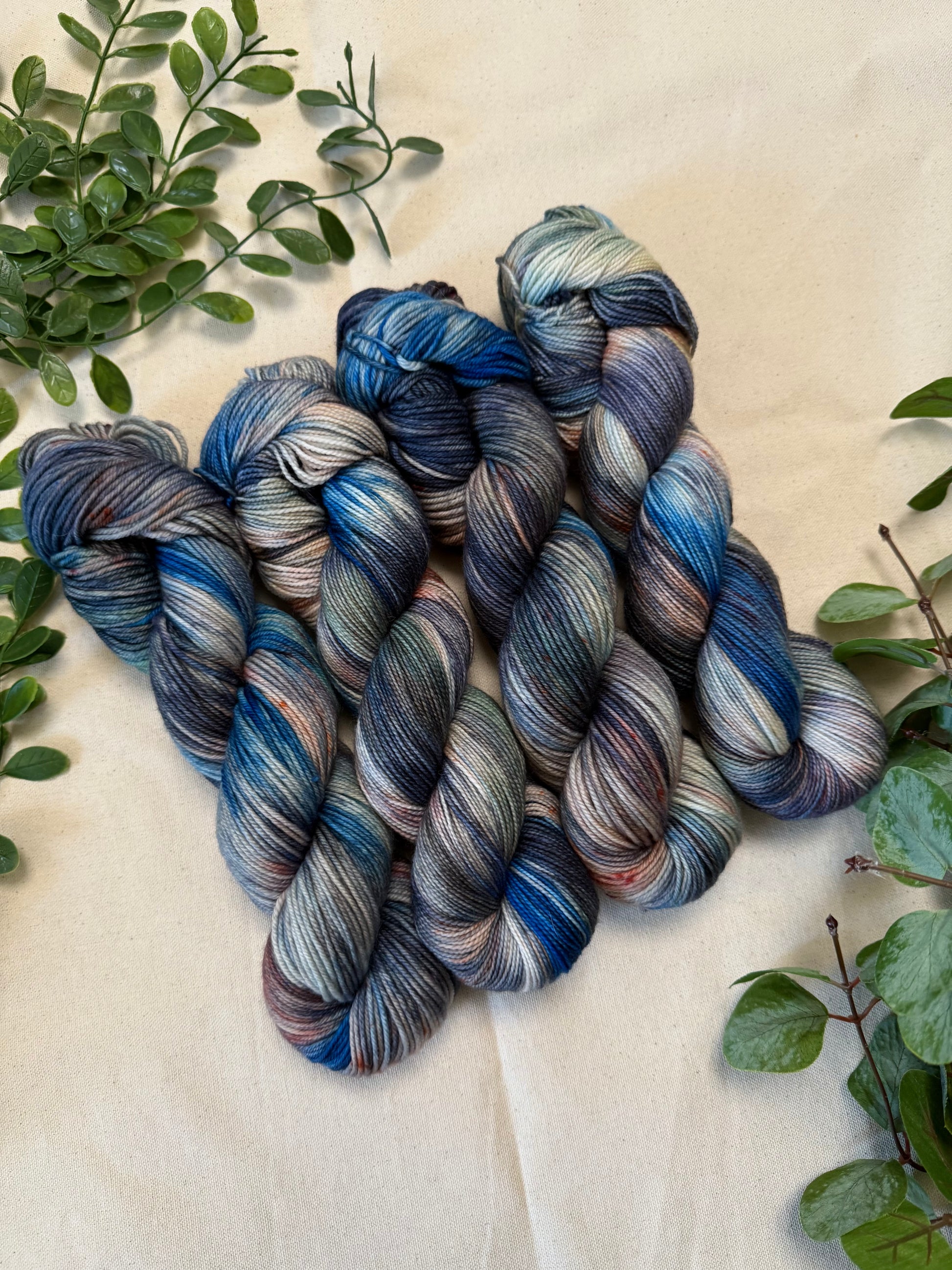 Four hanks of variegated yarn with blue, gray, and beige tones, arranged on a beige fabric background with green leaves.