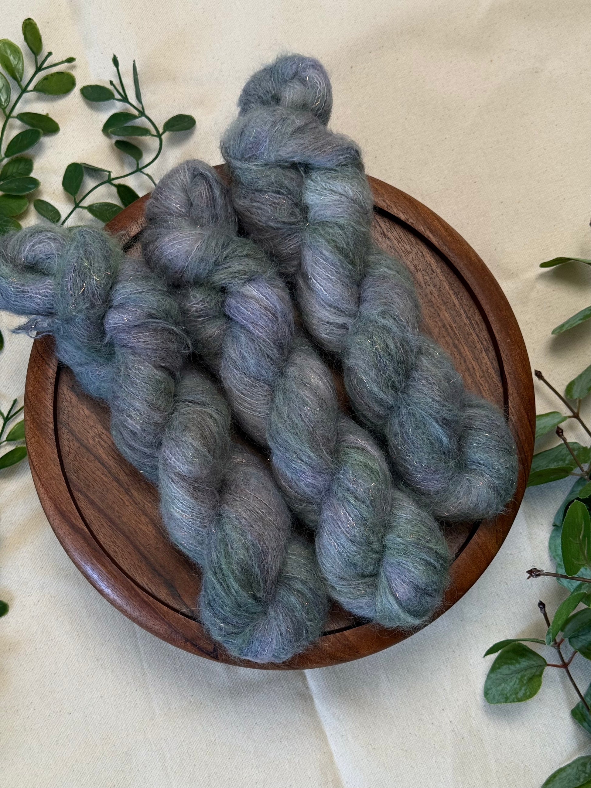 Three hanks of variegated blue-gray and green yarn displayed on a round wooden tray, surrounded by green leaves on a beige fabric background.
