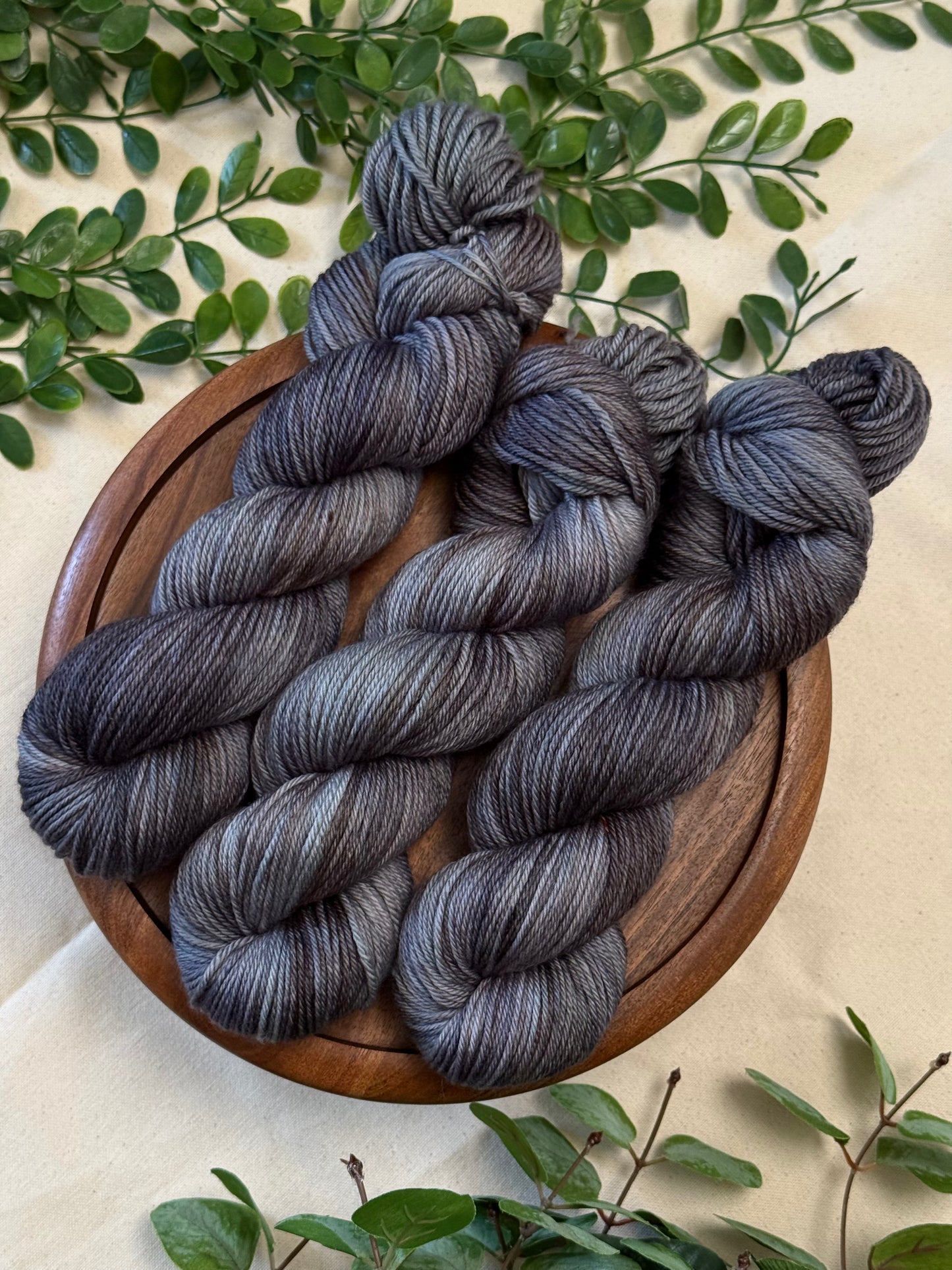 Three hanks of variegated gray and black yarn arranged on a round wooden plate, surrounded by green leaves on a light beige fabric background.
