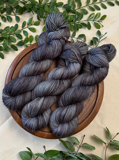 Three hanks of variegated gray and black yarn arranged on a round wooden plate, surrounded by green leaves on a light beige fabric background.