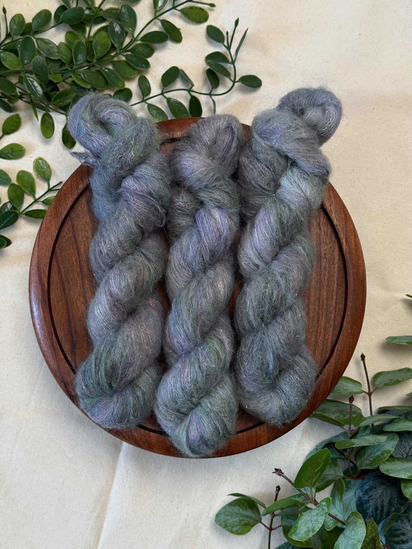 Three twisted hanks of fuzzy yarn in a mottled gray and lavender-blue shade, arranged on a round wooden tray with green leaves around it on a light fabric background.