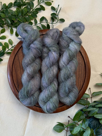 Three twisted hanks of fuzzy yarn in a mottled gray and lavender-blue shade, arranged on a round wooden tray with green leaves around it on a light fabric background.