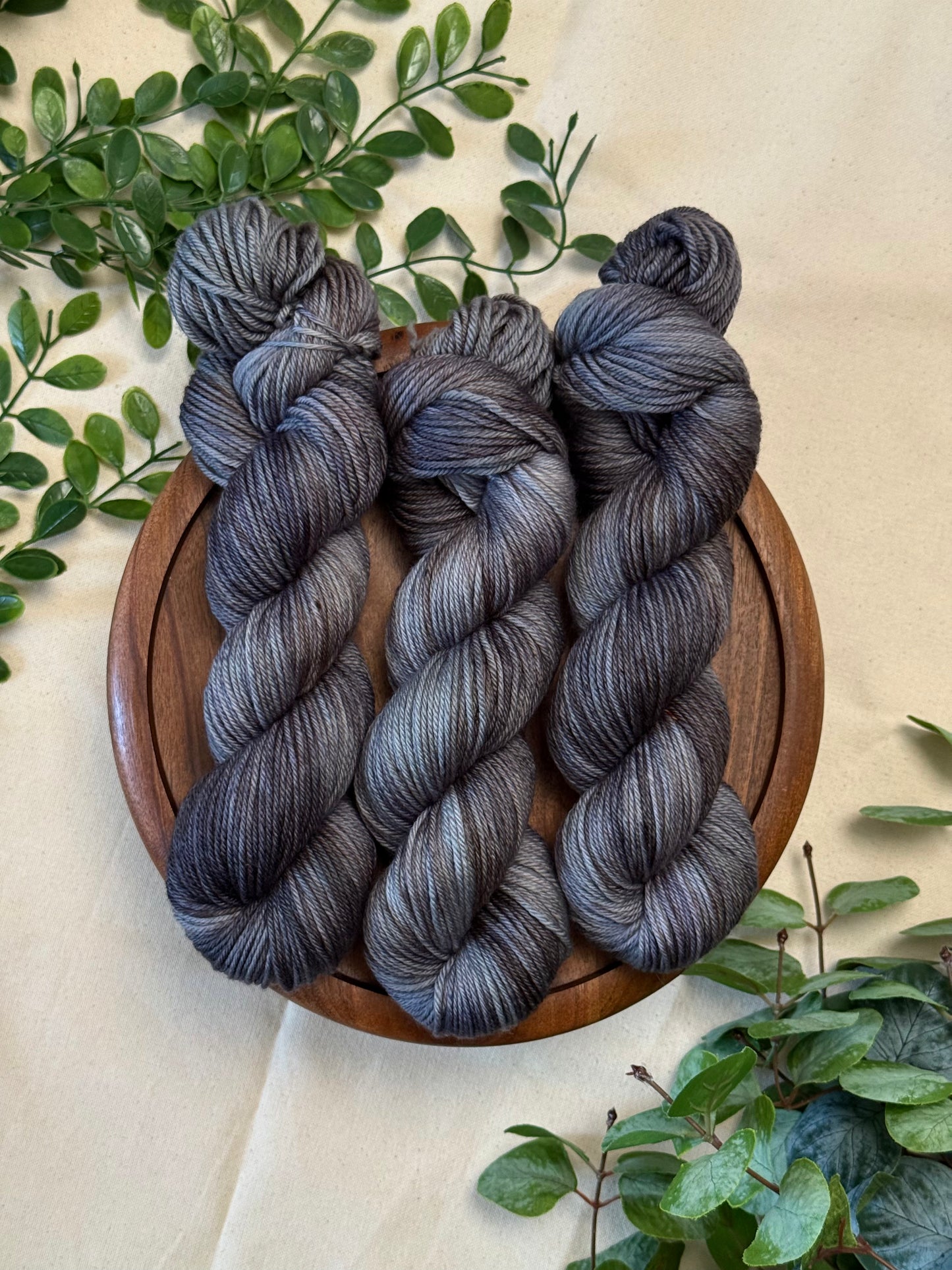 Three hanks of variegated gray and black yarn displayed on a round wooden tray, surrounded by green foliage on a beige fabric background.