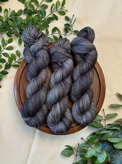 Three hanks of variegated gray and black yarn displayed on a round wooden tray, surrounded by green foliage on a beige fabric background.
