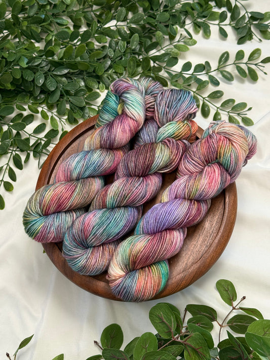 Four hanks of variegated merino wool yarn in pastel rainbow colors, arranged on a round wooden plate with green leaves on a white fabric background.