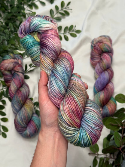 A hand holds a twisted hank of variegated yarn with pastel pink, blue, teal, and purple tones, with three more skeins arranged around it on a light fabric background with green leaves.