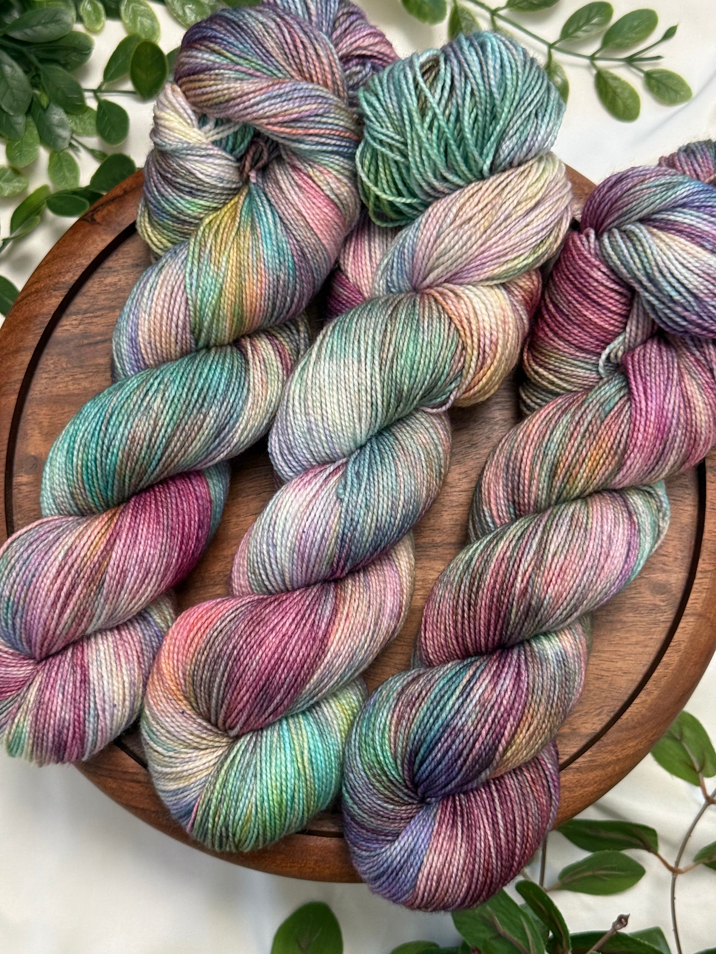 Four hanks of variegated merino wool yarn in pastel shades of teal, purple, pink, and green, arranged on a round wooden board with green leaves around the edges.