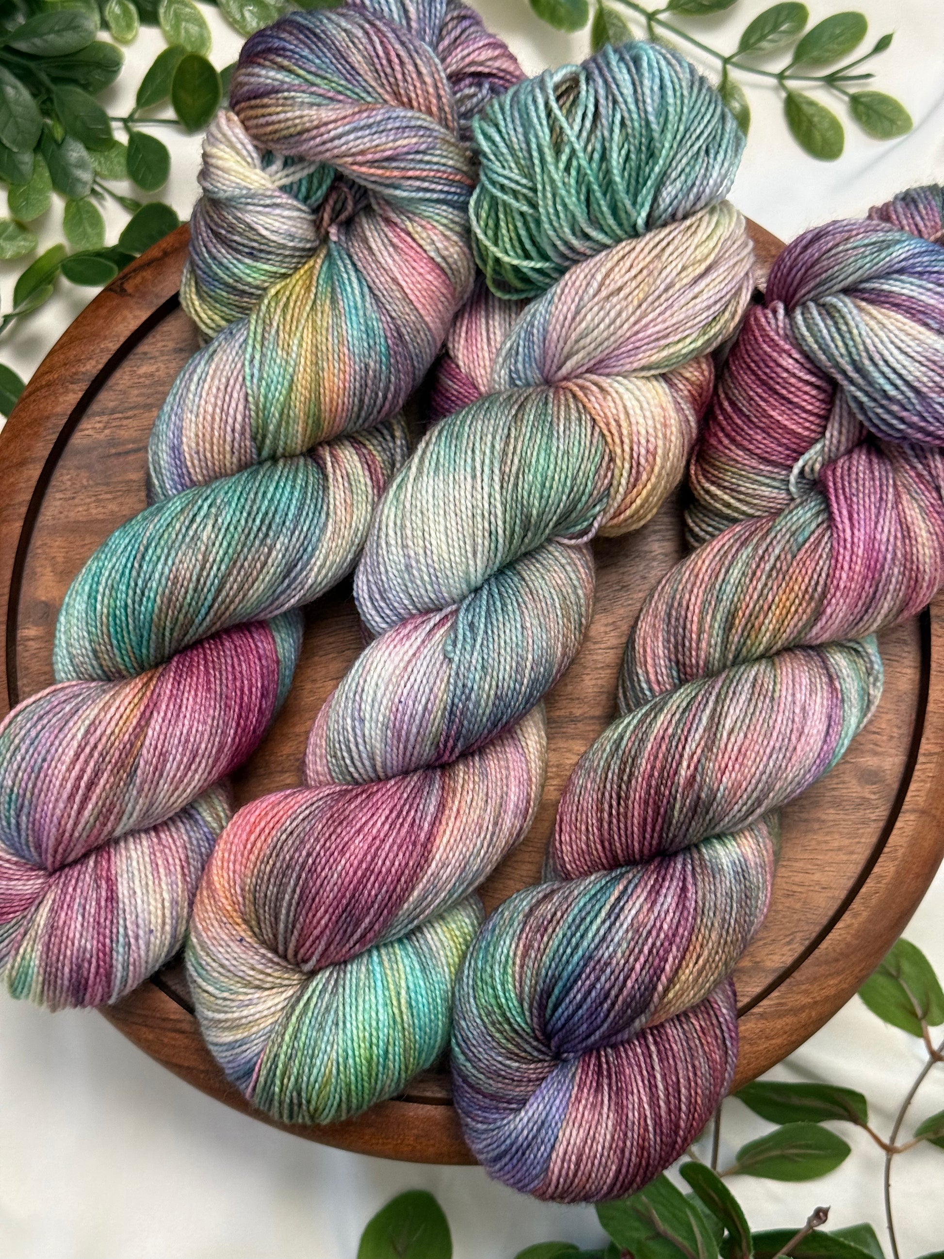 Four hanks of variegated merino wool yarn in pastel shades of teal, purple, pink, and green, arranged on a round wooden board with green leaves around the edges.