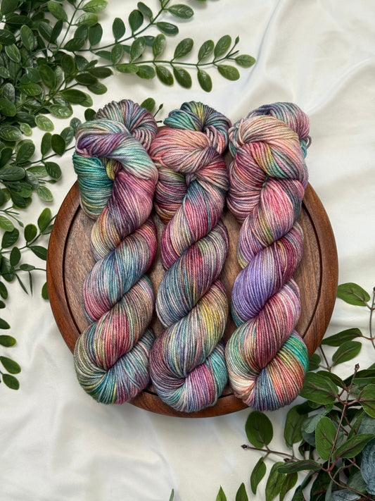 Three twisted hanks of variegated yarn in pastel rainbow shades, arranged on a round wooden tray over a white fabric background with green leaves around the edges.